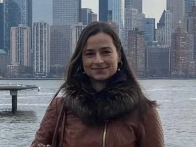 A woman in a brown coat with a fur collar standing by a railing on a waterfront, with the New York City skyline, including the One World Trade Center, in the background during cloudy weather.
