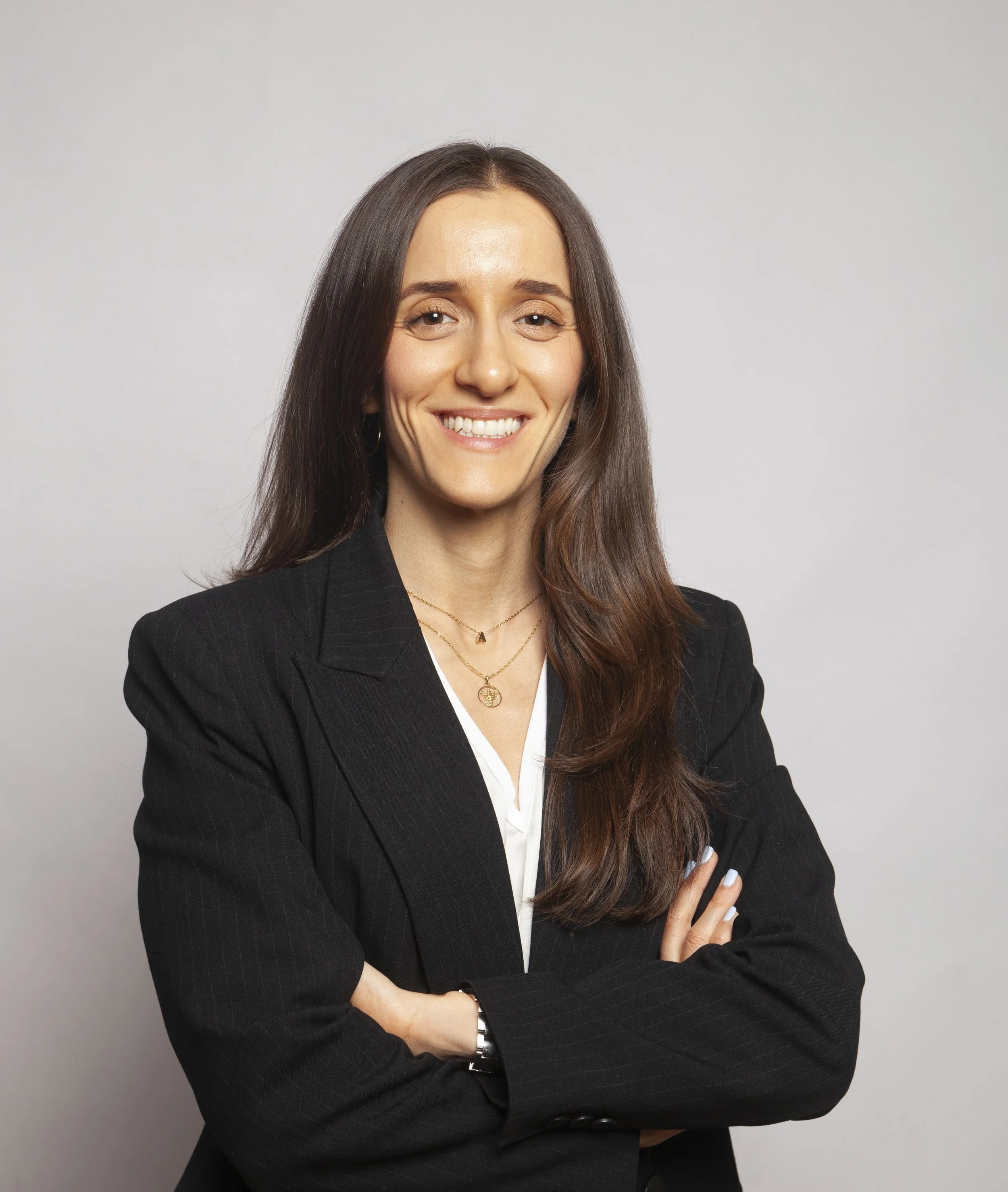 A woman with brown hair, smiling, dressed in a black blazer, standing against a plain light gray background.
