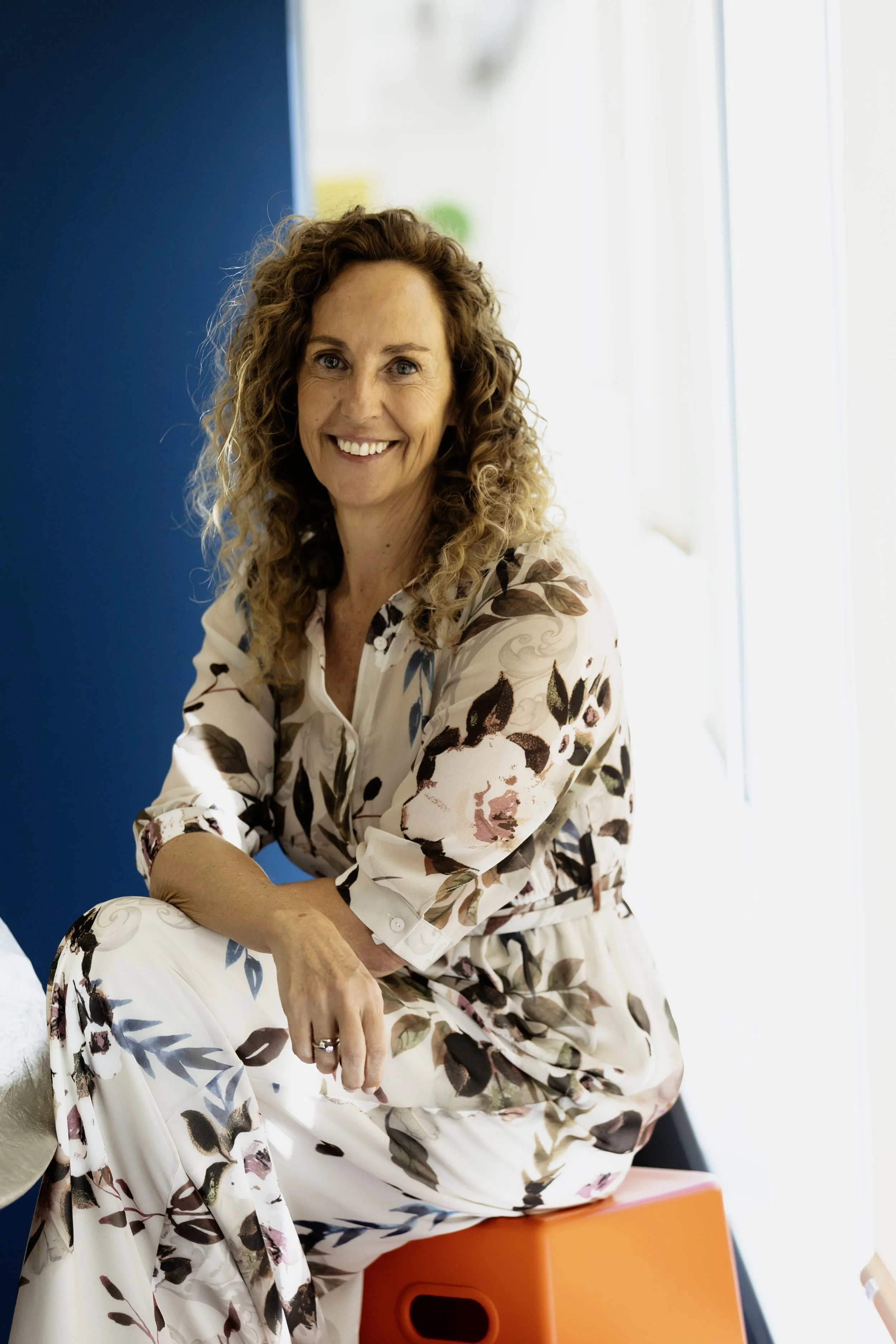 A woman with curly brown hair smiling and sitting on an orange object, wearing a floral outfit, in an indoor space with a blue wall and light background.