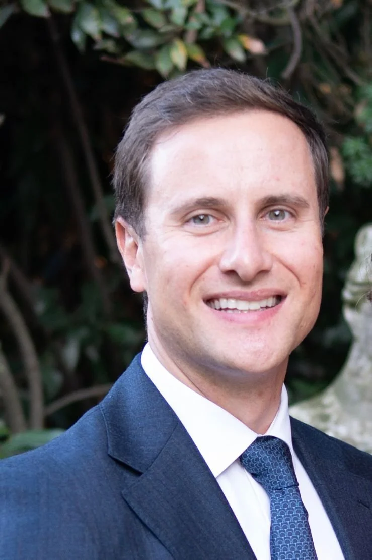 A smiling man in a dark suit, white shirt, and blue tie, standing outdoors with green foliage in the background.