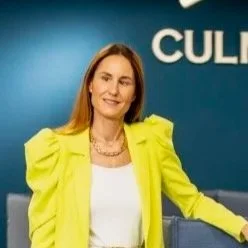 A woman with long brown hair wearing a bright yellow blazer and white top, standing in front of a blue background with the word 'CULT' partially visible.