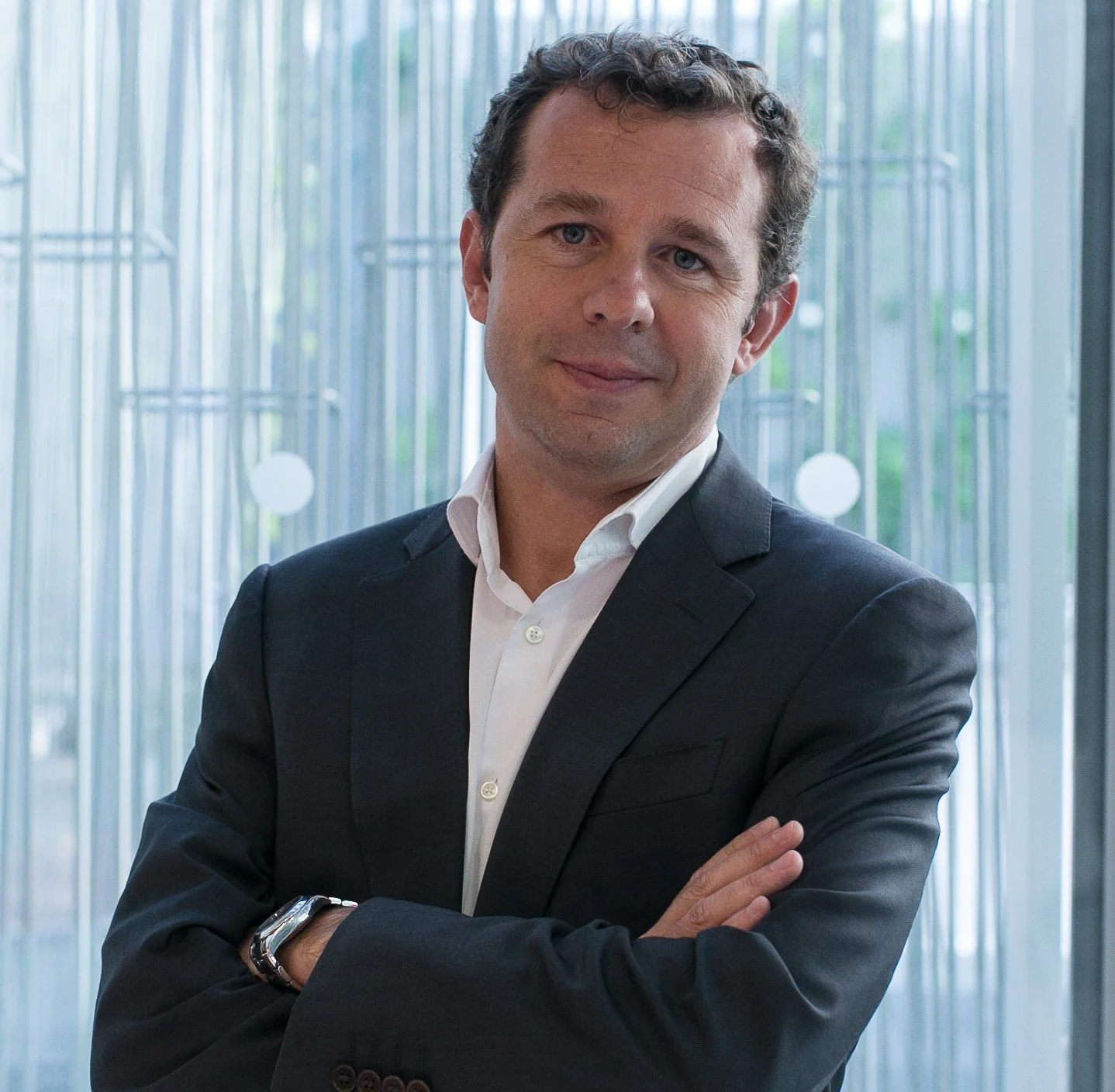 A man in a dark suit and white shirt standing with arms crossed in front of a glass wall with vertical lines and circular patterns.