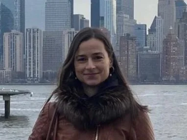 A woman standing by a waterfront with the New York City skyline in the background, including the One World Trade Center, on a cloudy day.