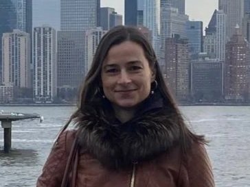 A woman in a brown coat standing by a railing along a waterfront with a city skyline featuring tall skyscrapers, including the One World Trade Center, in the background.