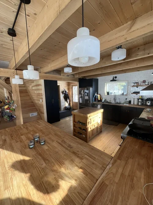 The image shows a cozy kitchen with wooden accents, including a wooden island and countertops, and exposed wooden beams on the ceiling. There are multiple white pendant lights hanging from the ceiling, a black cabinet, and various kitchen appliances 