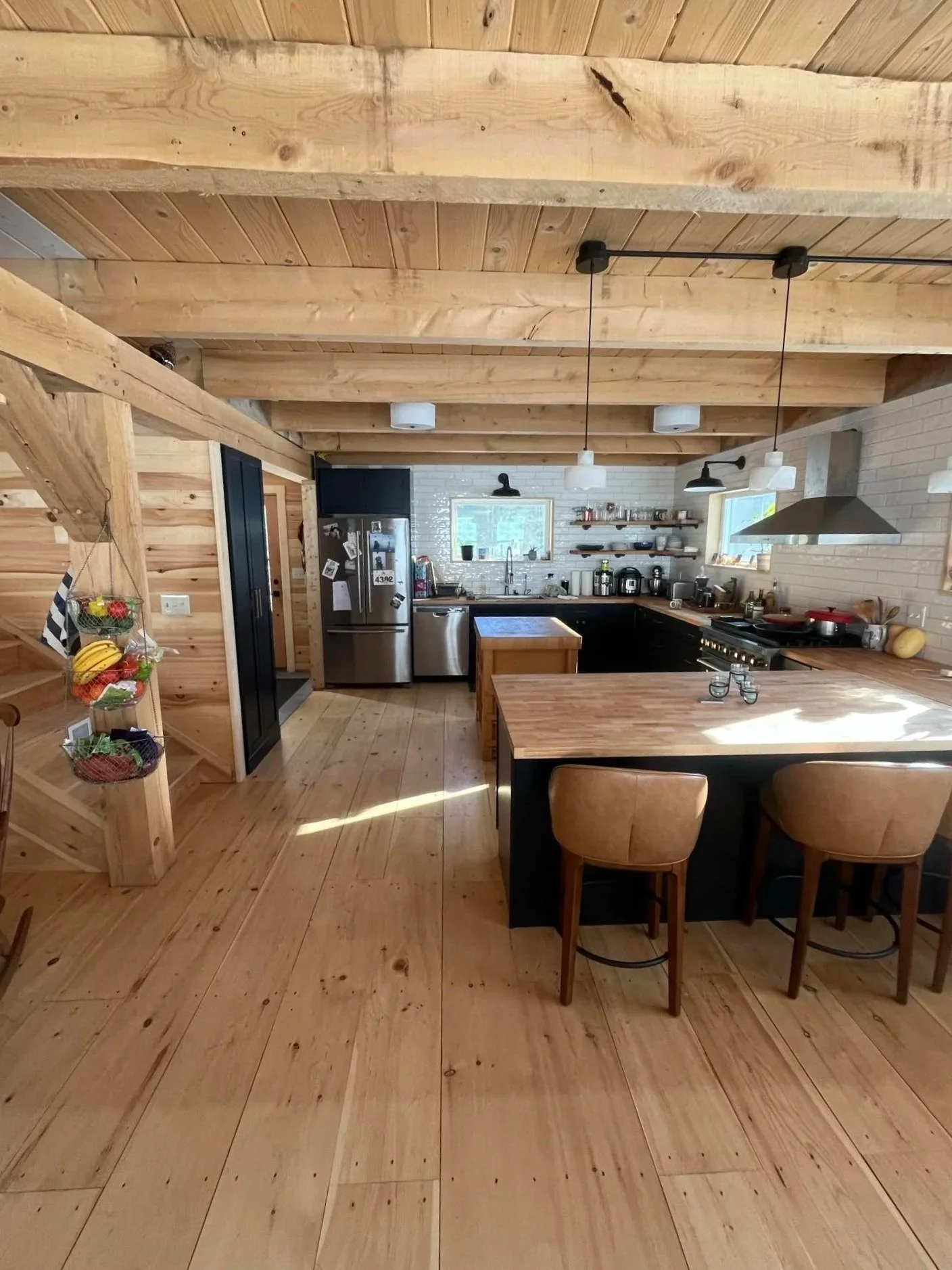 A rustic kitchen with wooden floors, ceiling, and walls, including a central kitchen island, stainless steel appliances, open shelves with dishes, and a breakfast bar with stools.