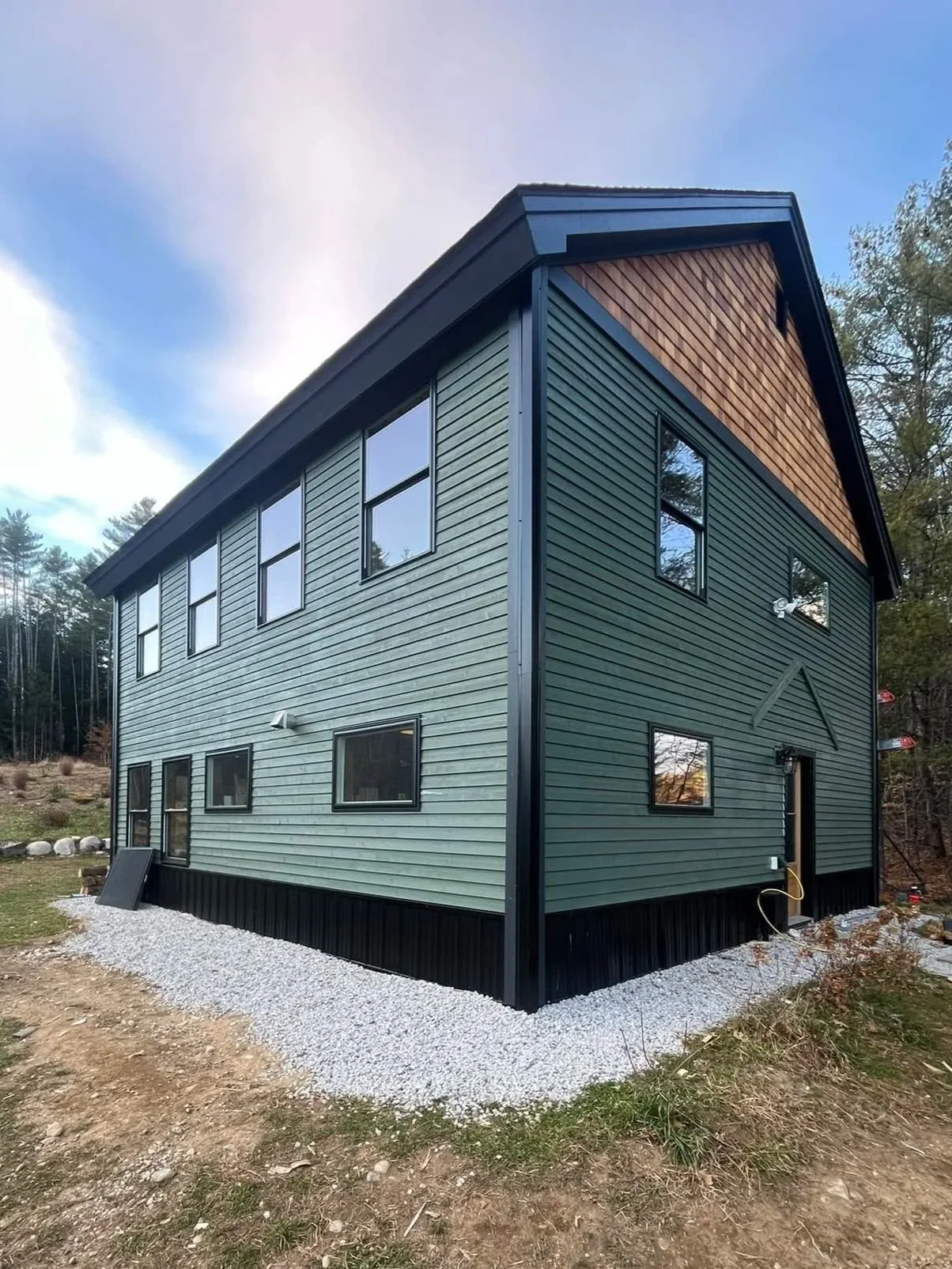 A modern two-story house with green siding, black trim, multiple windows, and a wooden panel accent on the gable end, set on a gravel base outdoors.