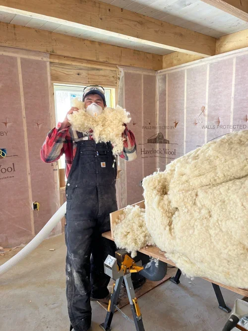 A construction worker wearing a dust mask and protective glasses in an unfinished room, holding a large piece of insulation material.