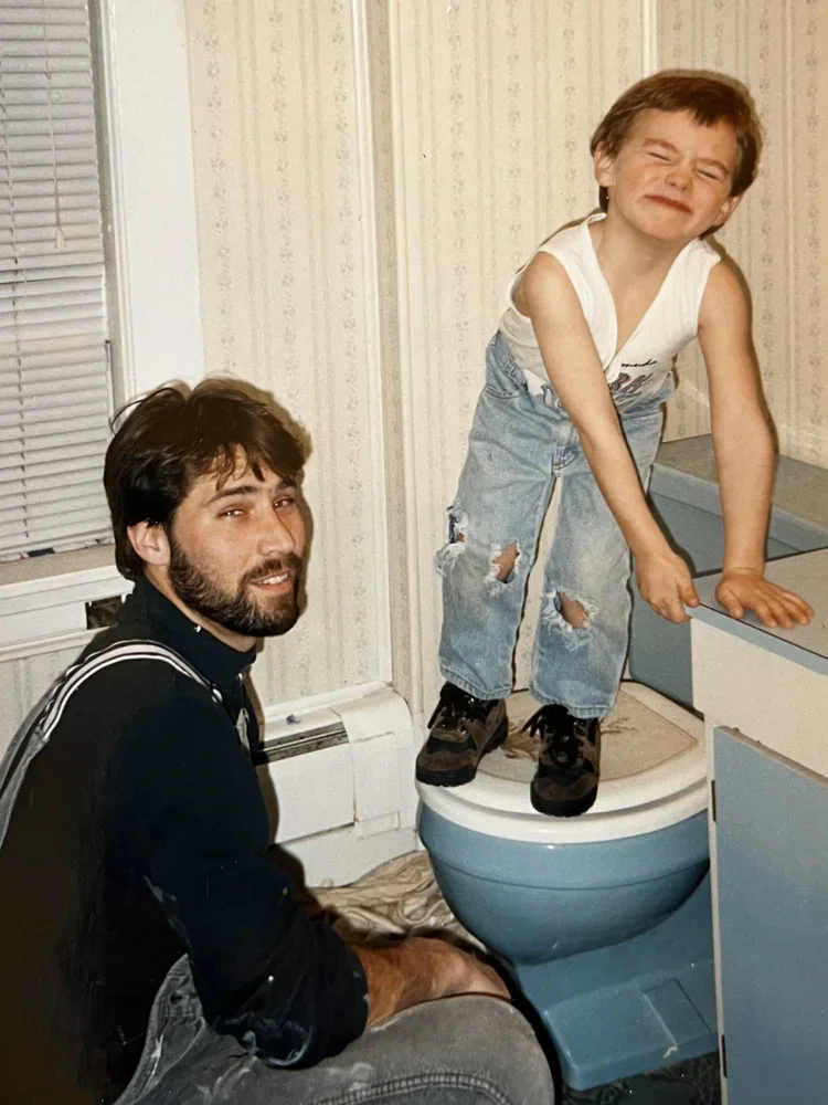 A young boy with short brown hair, wearing a white sleeveless shirt and ripped jeans, standing on a toilet seat and smiling with eyes closed, next to a man with dark hair and beard, wearing a black shirt with suspenders, sitting on the floor in a bathroom with floral wallpaper and a window with blinds.