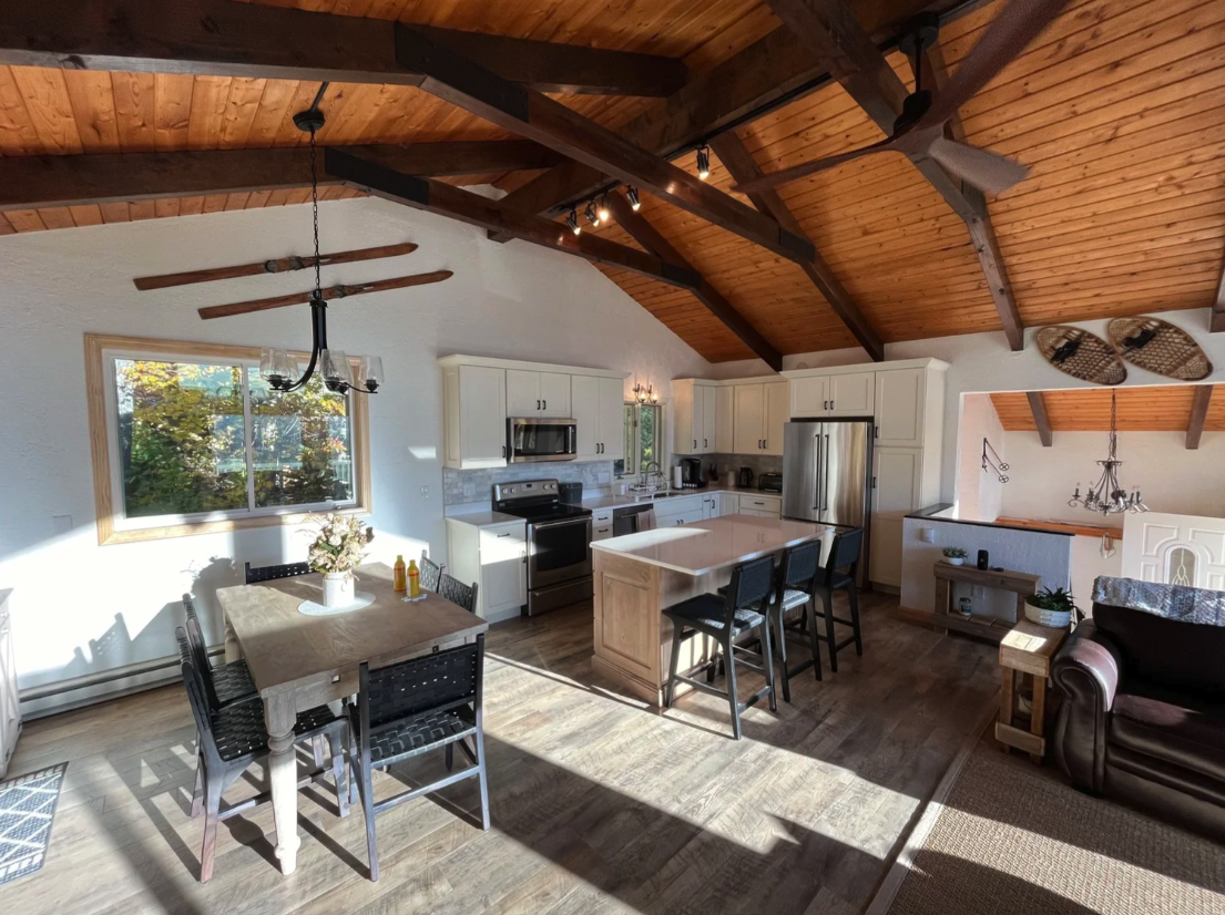 Open concept kitchen and dining area with wooden vaulted ceiling, white cabinets, stainless steel appliances, and a kitchen island with barstools. A wooden dining table with six chairs and a unique chandelier hang above it. Large window lets in natural light, with a view of trees outside.