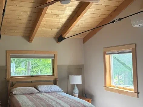 A bedroom with a wooden vaulted ceiling, a bed with gray and white bedding, a window above the bed with a view of greenery outside, a bedside table with a white lamp, and another window with a view of trees outside.