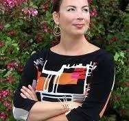 A woman standing outdoors in front of a flowering bush, wearing a black and white patterned top, with her arms crossed.