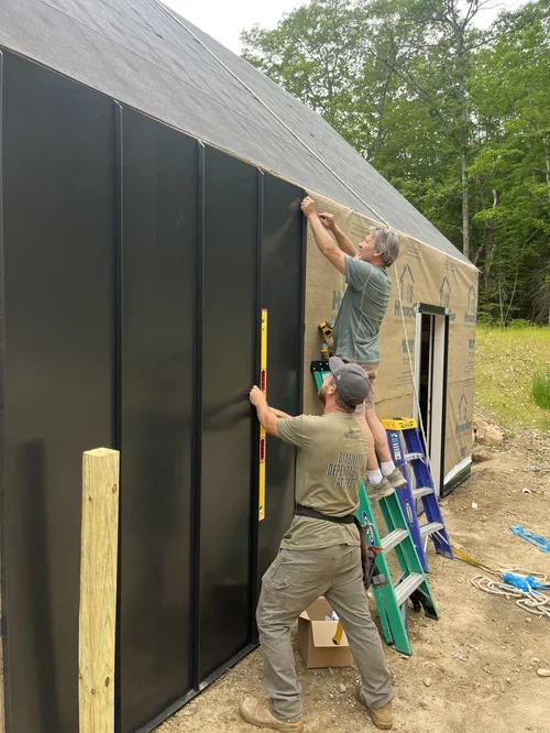 Two workers installing black wall panels on a building under construction in an outdoor setting.
