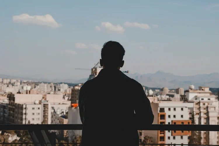 Person facing away, standing on a balcony overlooking a city skyline with mountains in the background.