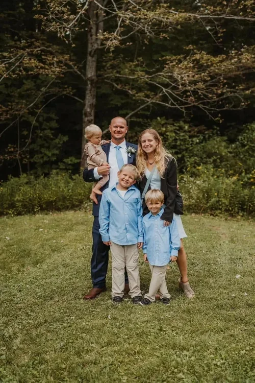 Family portrait of five people standing outdoors on a grassy area with trees in the background.