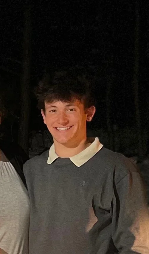 A young man with dark, curly hair smiling at the camera, dressed in a dark gray shirt with a white collar, standing outdoors at night.