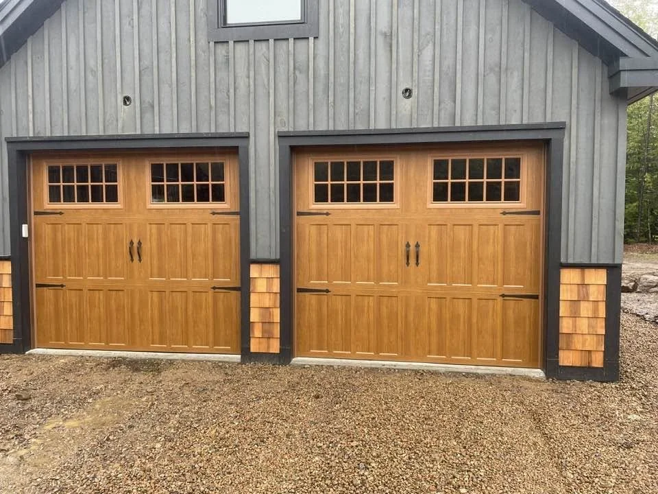 Double garage doors with small windows at the top and black handles, attached to a building with gray vertical siding and decorative wood panels beneath the windows, on a gravel driveway.