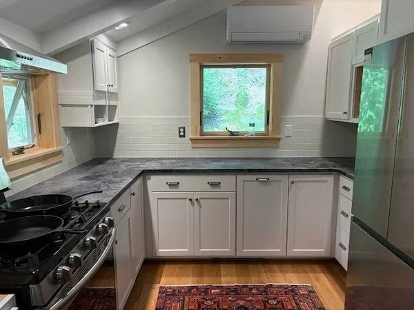 Kitchen with white cabinets, dark granite countertops, a window with green trees outside, a stainless steel refrigerator, an oven with multiple burners, and a red patterned rug on a wooden floor.