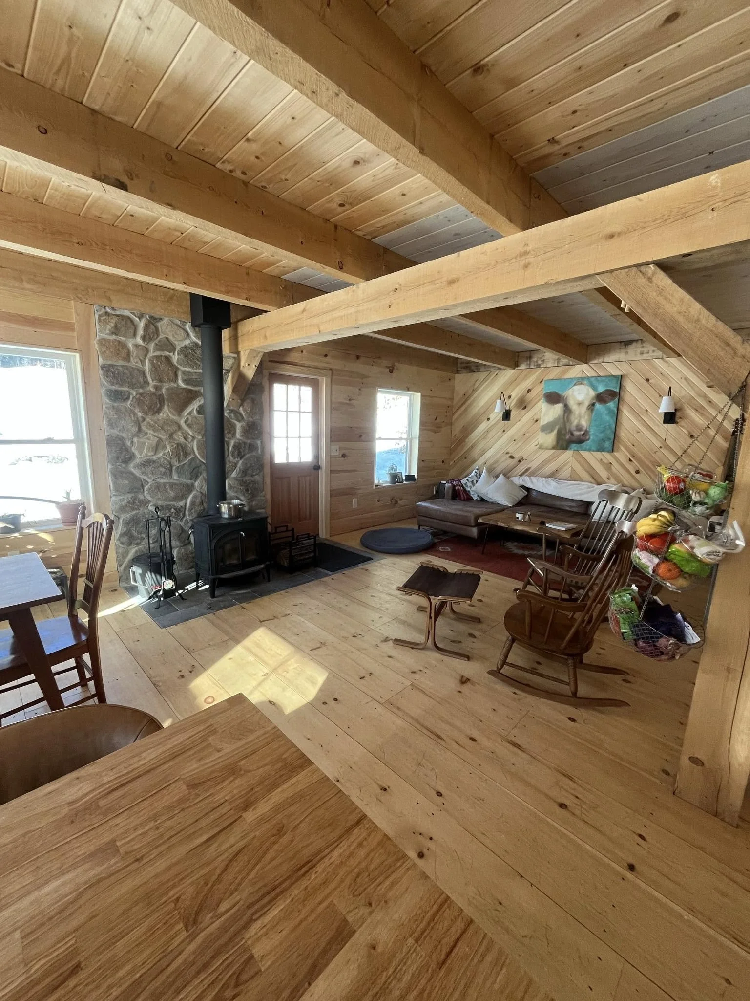 Inside a wooden cabin living room with high vaulted ceiling, exposed wooden beams, wood-paneled walls, a stone fireplace, a wood stove, a wooden table, chairs, a corner with a couch, a painting of a cow, and a window allowing natural light.