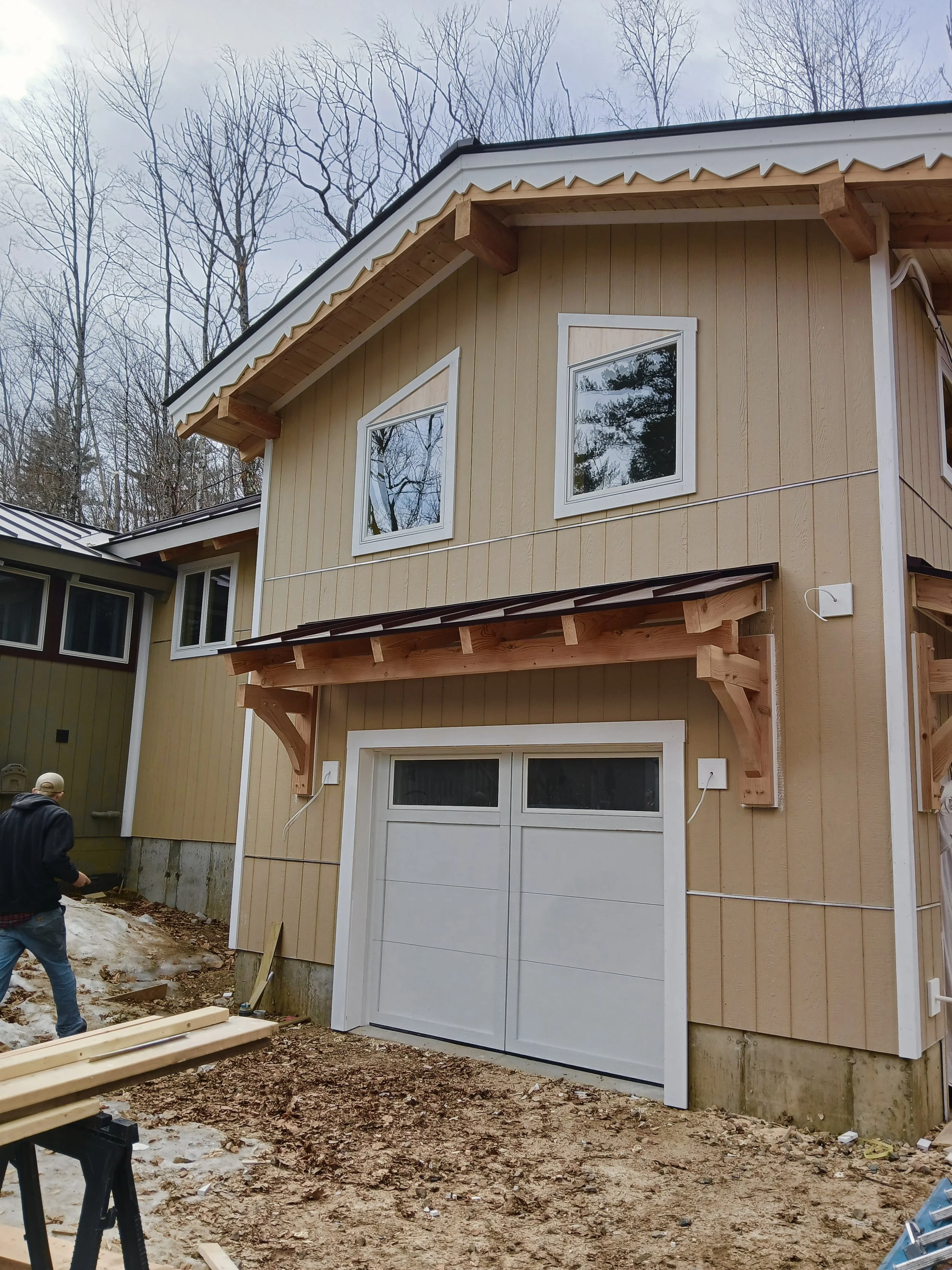 A house under construction with a new garage door installed. The house has beige siding, white window frames, and a wooden awning over the garage. The construction site has dirt, construction materials, and a person working.