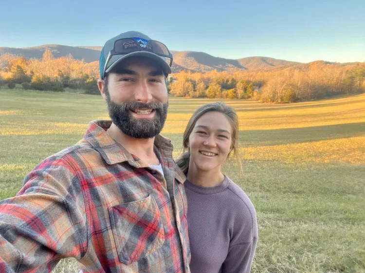 A man and woman smiling outdoors in a grassy field with mountains and trees in the background during sunset.