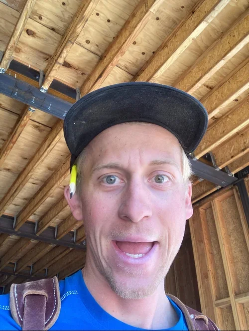 Man taking a selfie inside a wooden structure, wearing a baseball cap, a blue shirt, and a harness, with exposed wooden beams and insulation in the background.