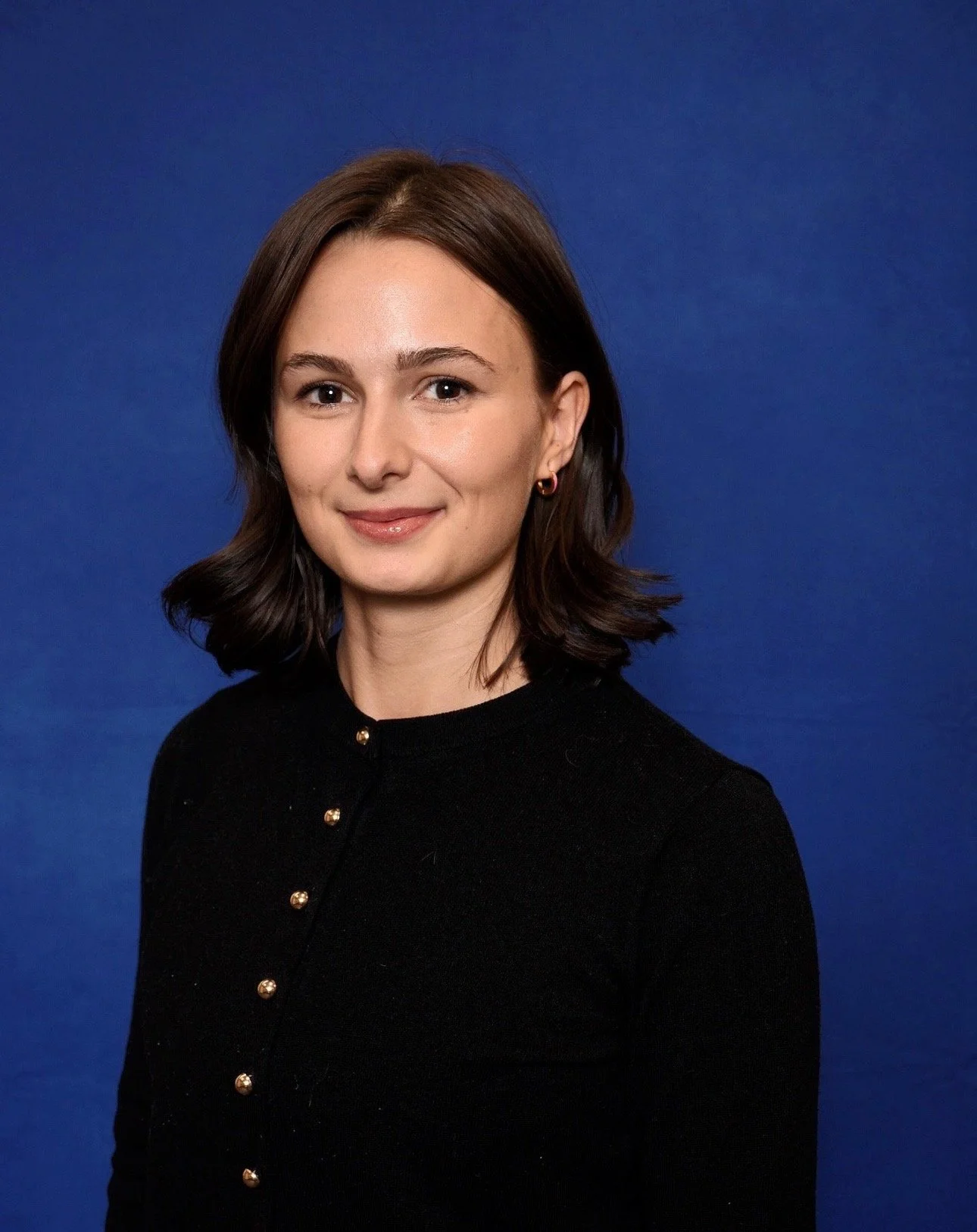 A woman with medium-length dark brown hair, fair skin, and blue eyes, smiling gently, wearing a black top with gold buttons, standing against a solid blue background.