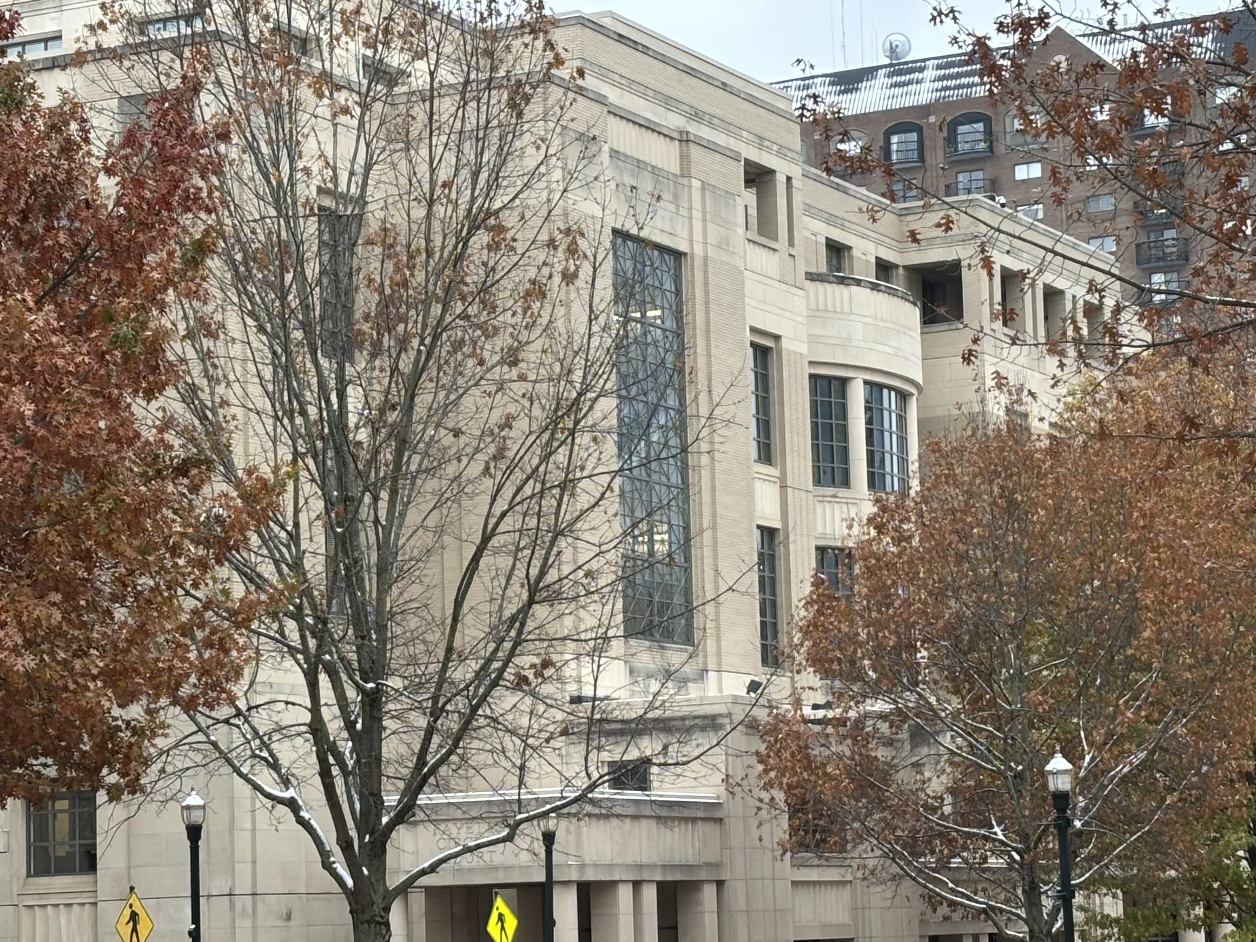 A city building with large windows and architectural details, partially obscured by trees with orange leaves, and sidewalk pedestrian signs at the bottom of the image.