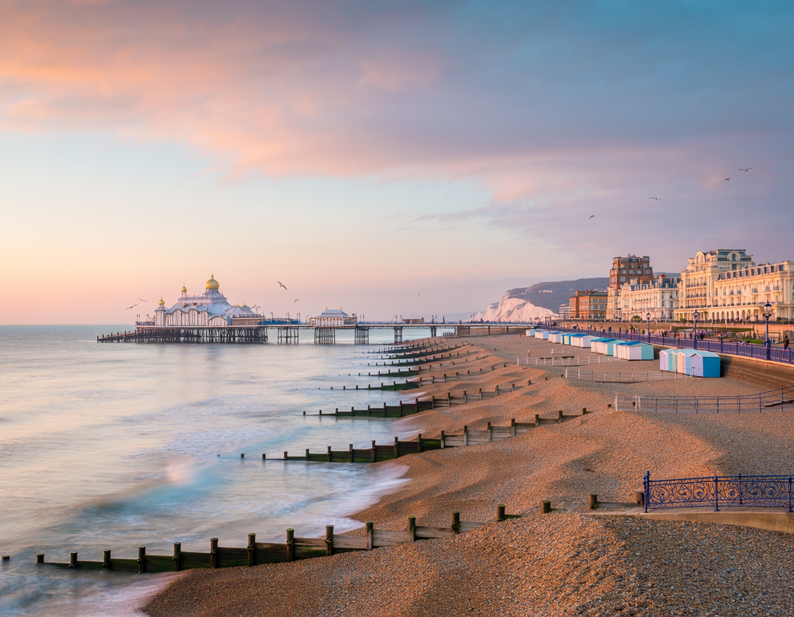View of a beach with wooden groynes, colorful beach huts, a pier with a pavilion at the end, and buildings along the coast at sunset.