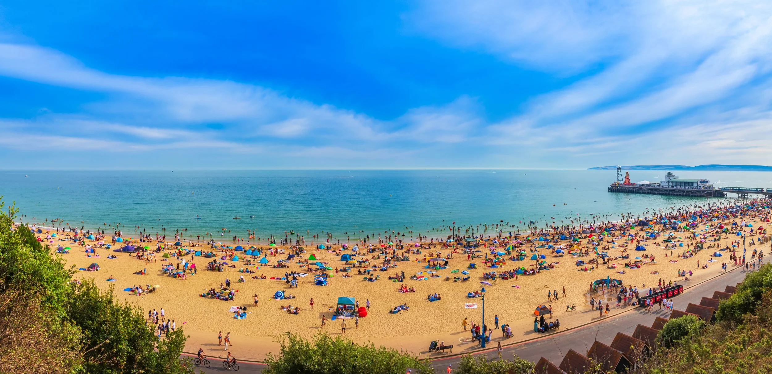 A busy beach scene with numerous people sunbathing, swimming, and enjoying the ocean. The sandy shore is filled with colorful umbrellas and towels. There is a pier extending into the water with a small building or attraction at the end. The sky is partly cloudy with a bright, sunny atmosphere.