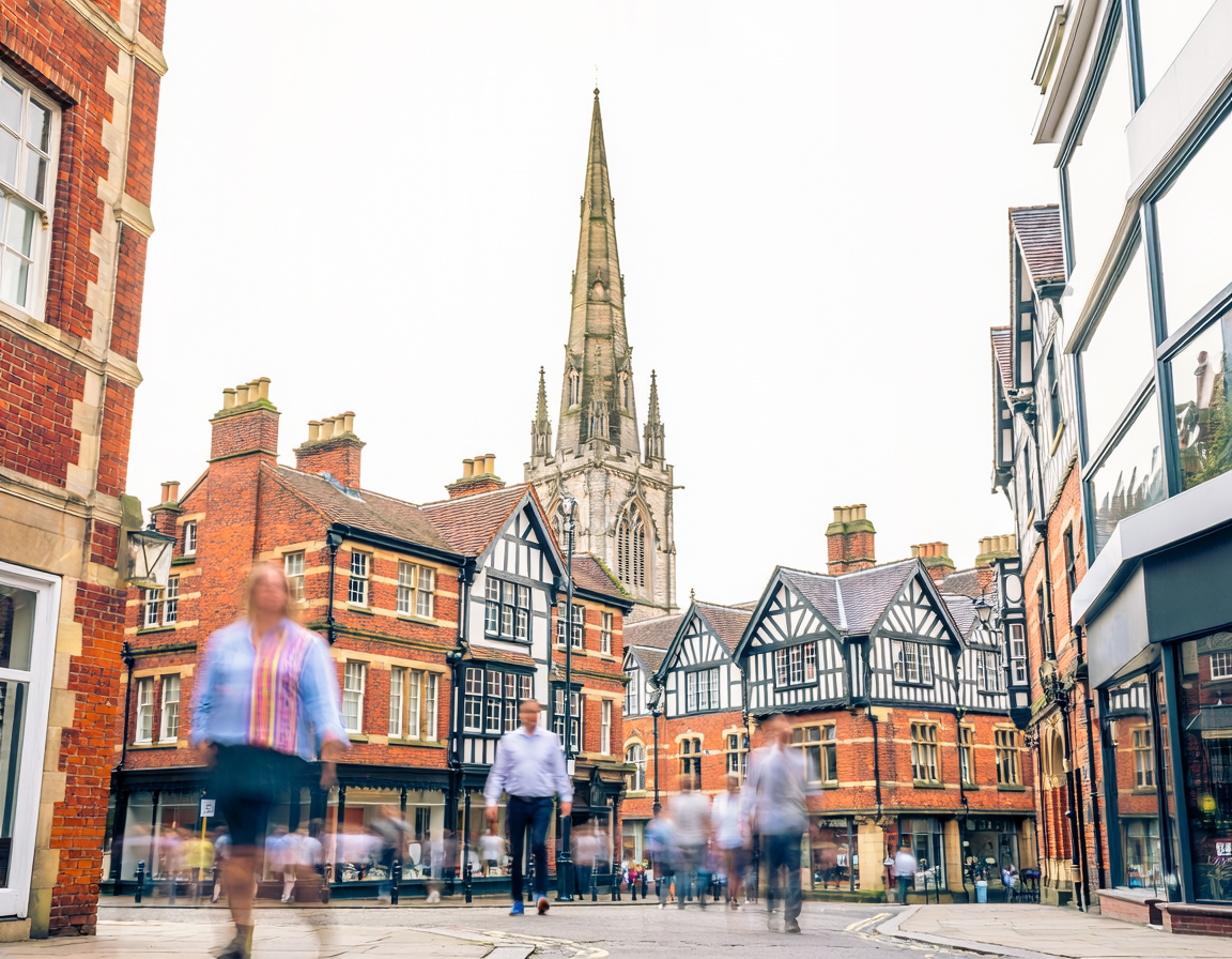 People walking in a city square with historic brick and timber-framed buildings, and a tall church steeple in the background.