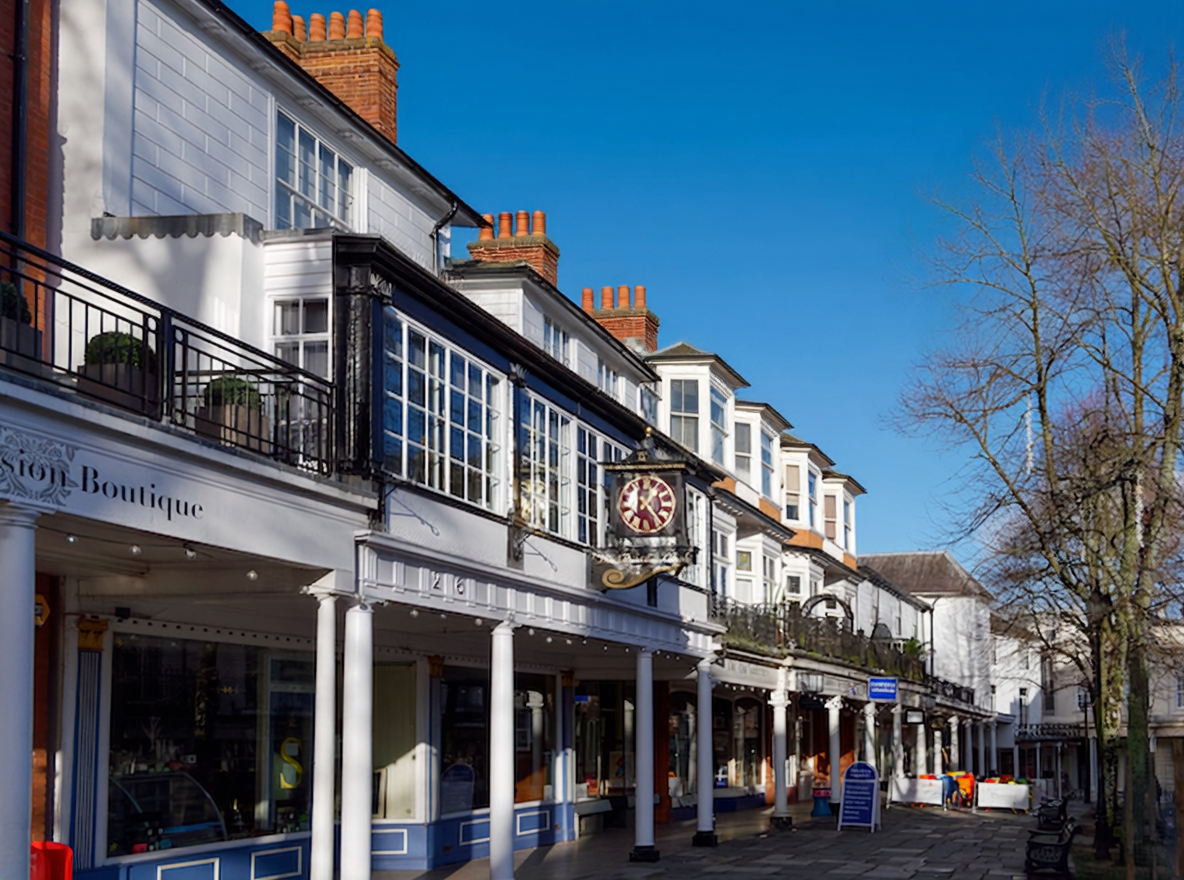 A row of white historic buildings with large windows and black iron balconies on a sunny day, with a sidewalk below and a leafless tree on the right.