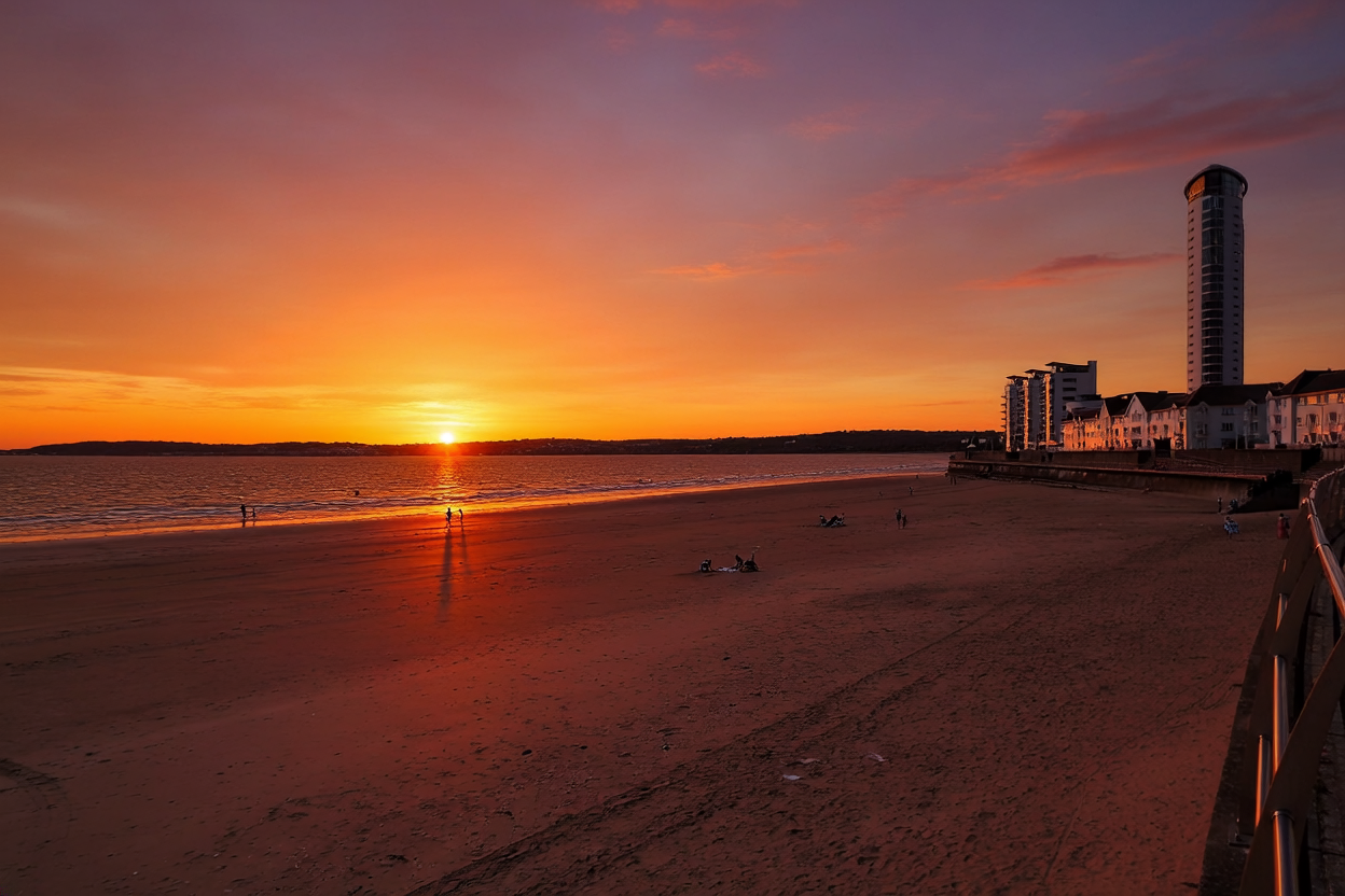 Sunset over a beach with a few people near the water, some sitting on the sand, and modern buildings including a tall tower along the shoreline.