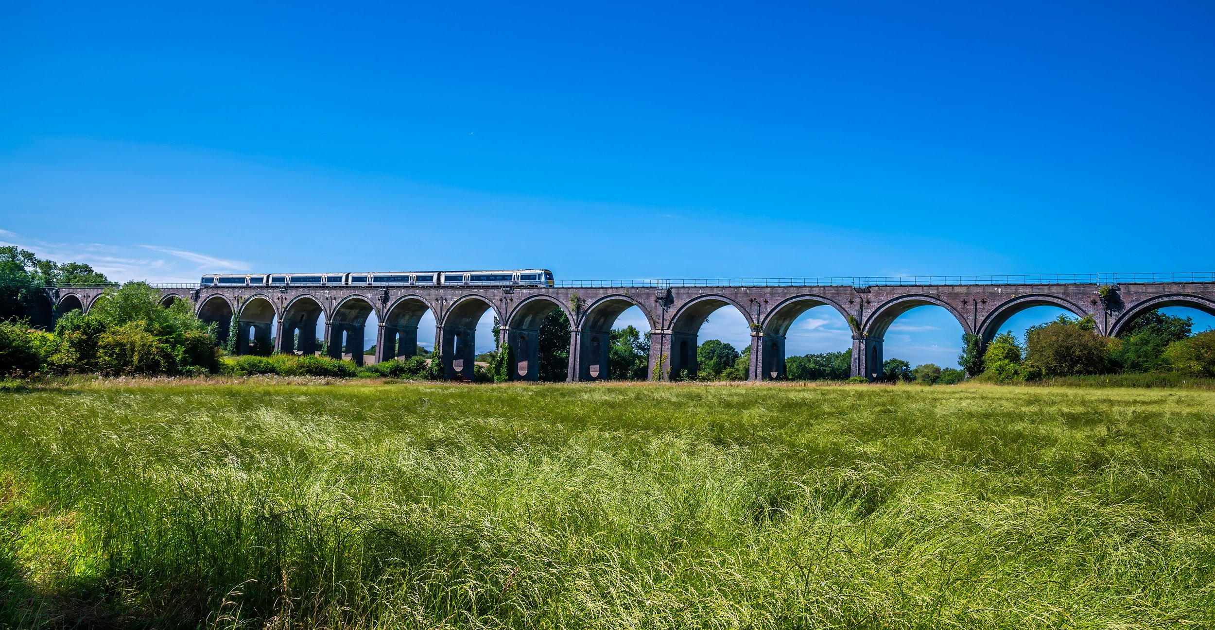 A train traveling over a large stone bridge with multiple arches, set against a bright blue sky and surrounded by greenery.