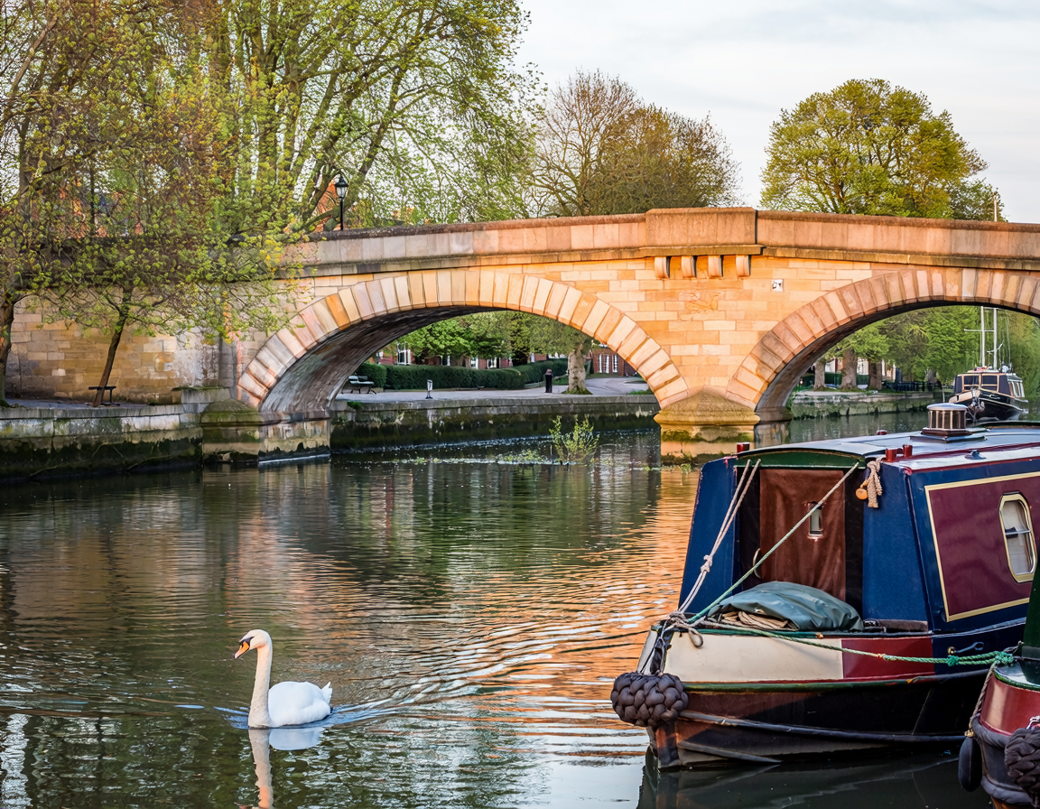 A scene featuring a stone bridge over a canal with boats, a swan swimming in the water, and trees with green and some orange leaves, indicating early spring or fall.