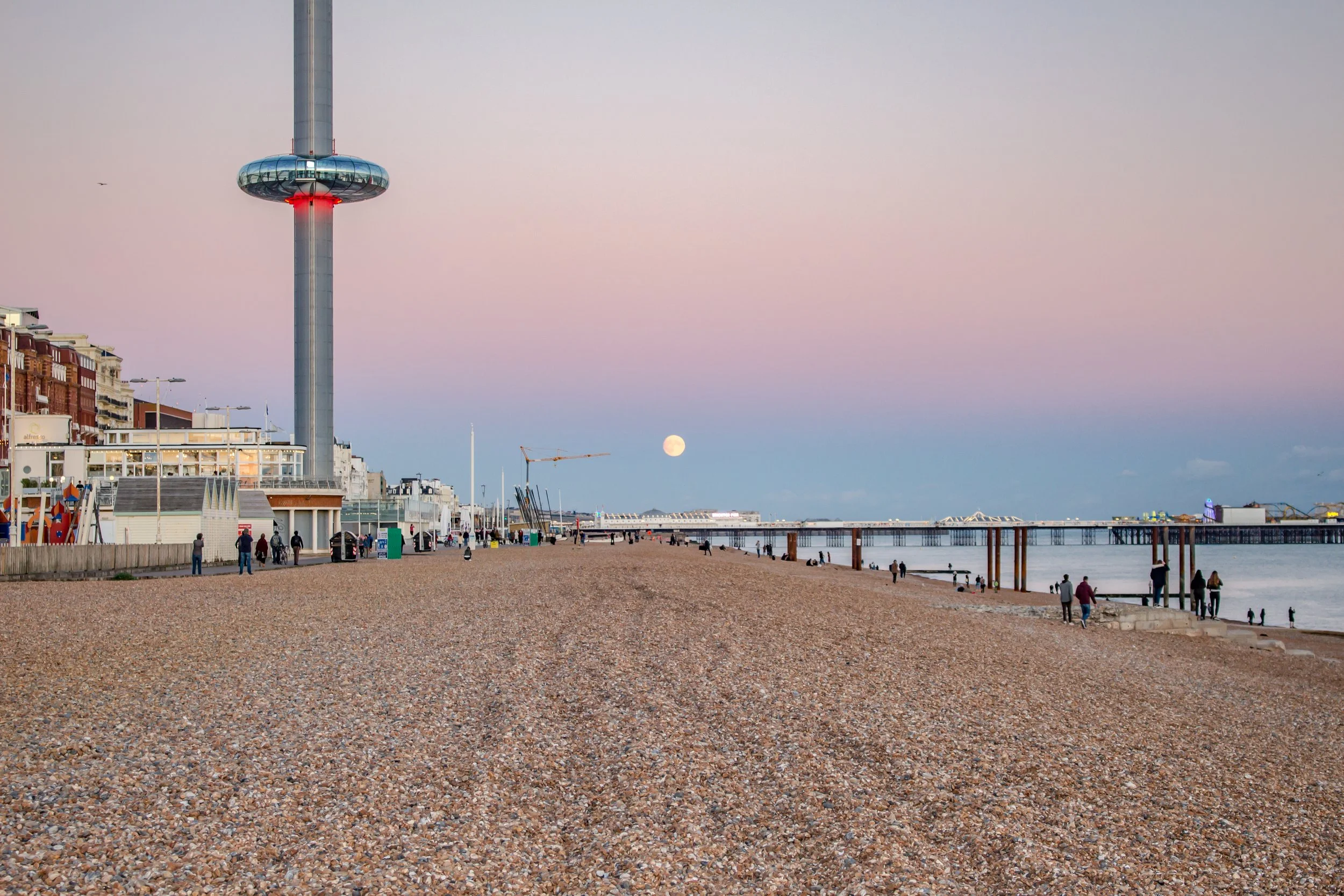 Beach with a pebbly shore, people walking, a pier, and an iconic tower with a circular observation deck, a red light, and a moon in the sky at dusk.