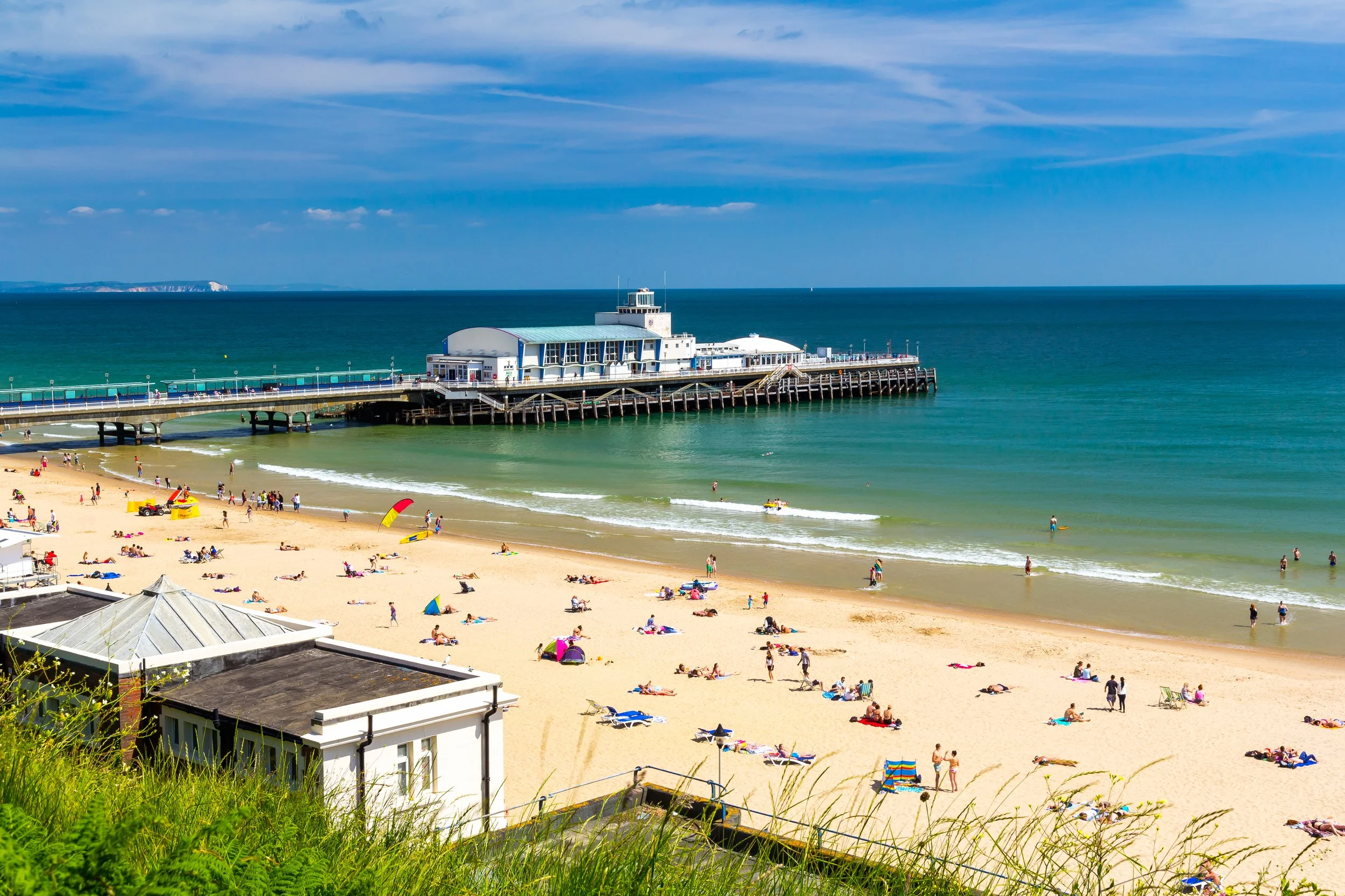 Beach with people sunbathing and swimming, pier with buildings extending into the water, blue sky with clouds, ocean waves, and a distant shoreline.