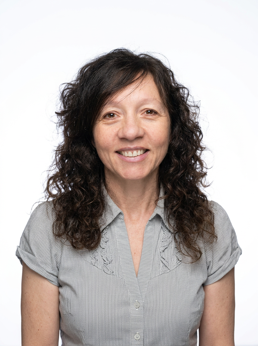 A woman with curly brown hair and a light gray short-sleeve blouse, smiling, against a plain white background.