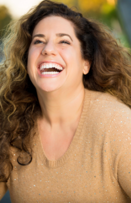 A woman laughing outdoors in a sunny setting with green foliage in the background.