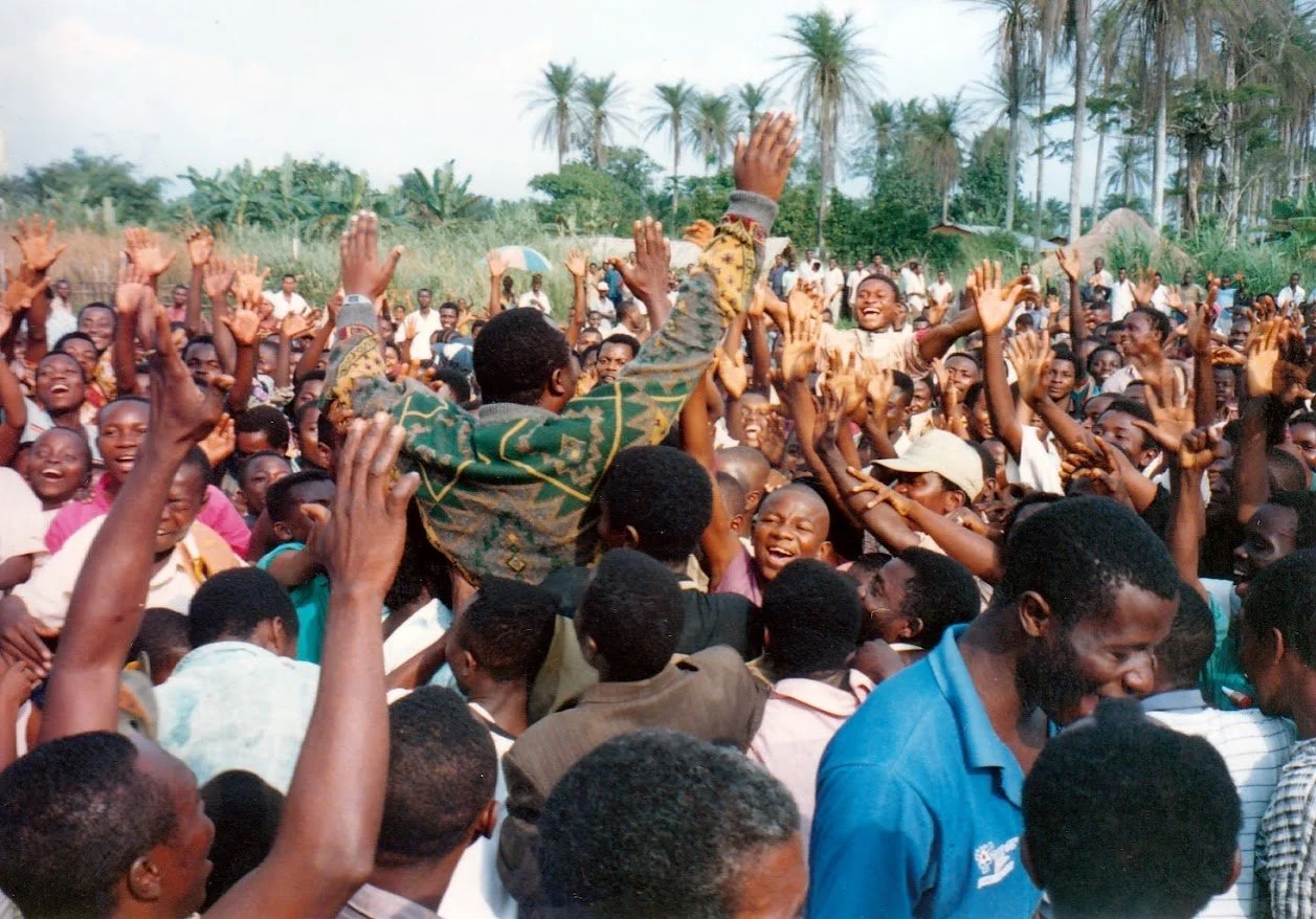 Group of people celebrating at airport of Shabunda, the arrival of Prof. Ambrose Walemba, some with raised hands, amidst trees and grassy area.