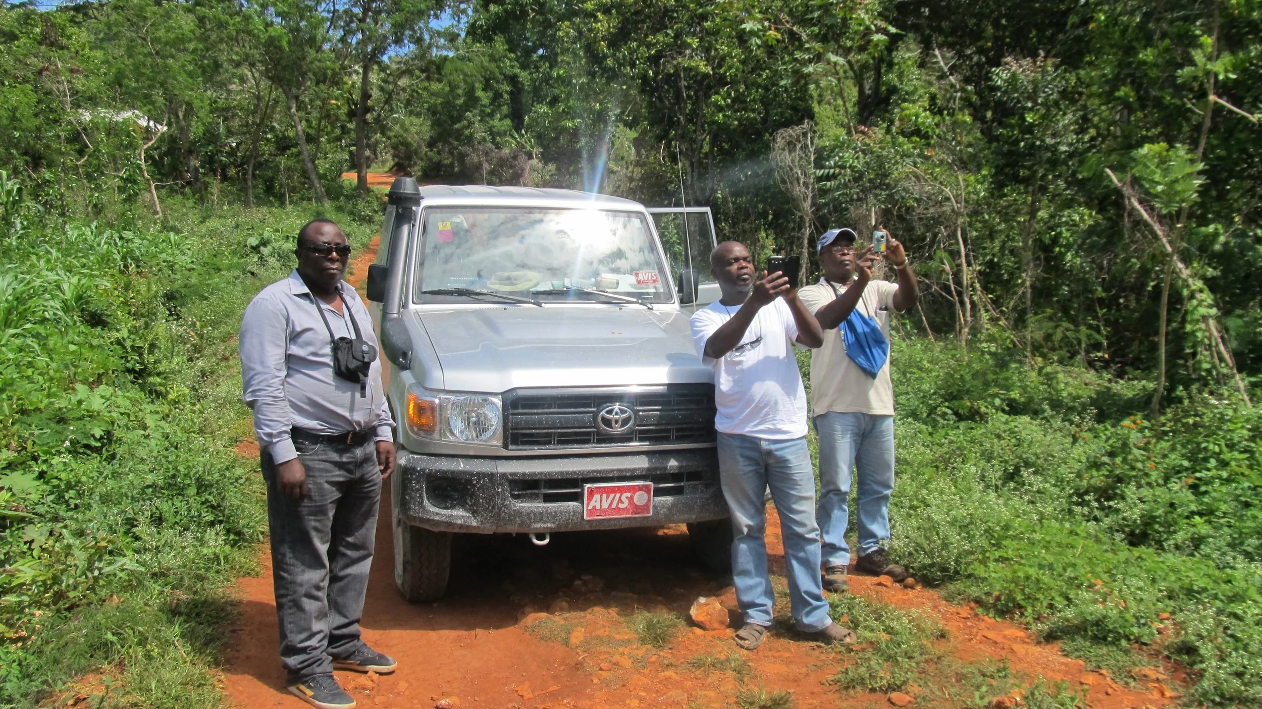Four men, including Prof. Ambrose Walemba, standing beside a gray Toyota vehicle on a dirt road surrounded by green trees and vegetation, with two of them taking photos or videos using their phones in Haiti.