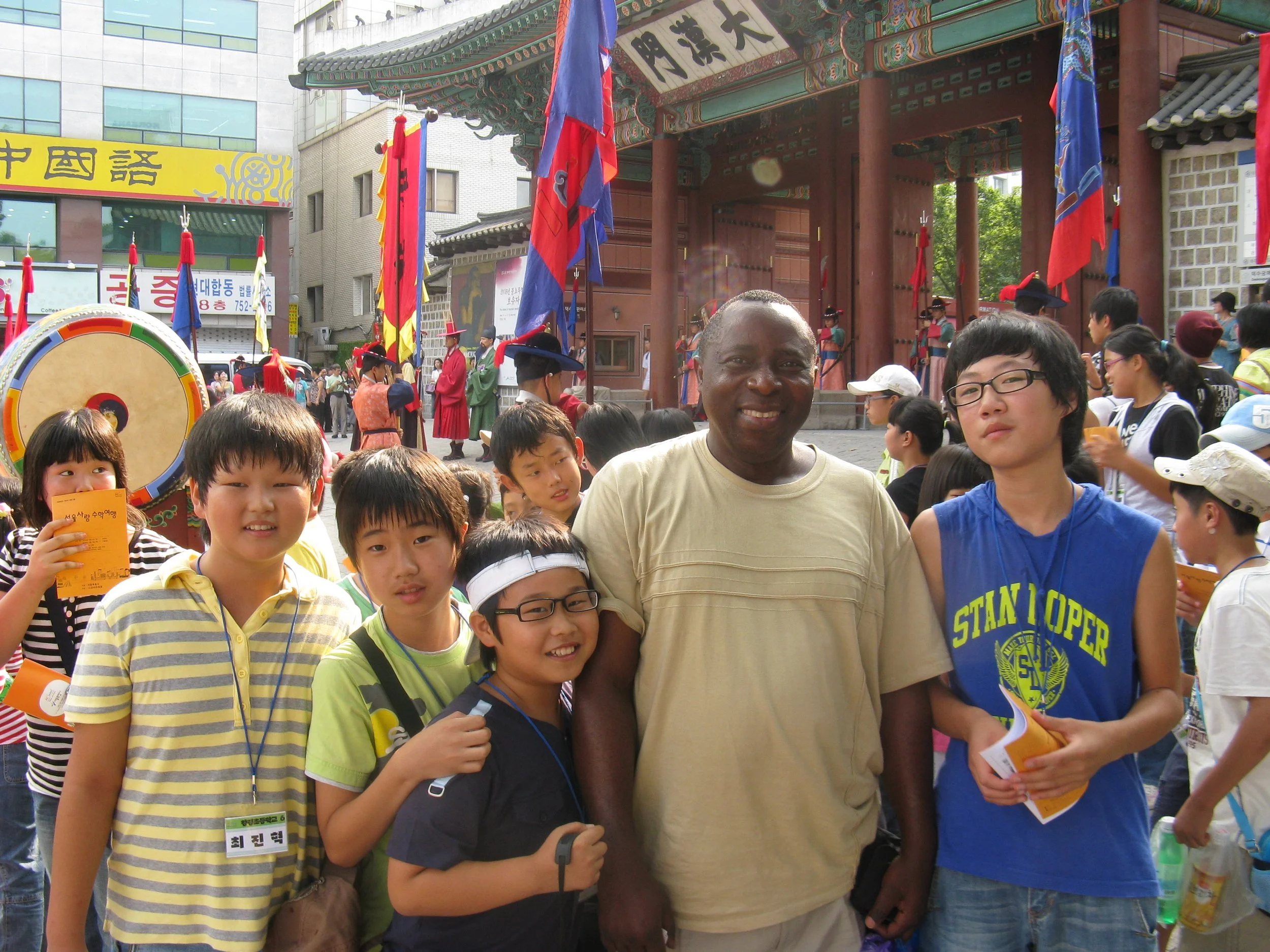 A diverse group of children and Prof. Ambrose Walemba are gathered in front of a traditional Korean gate during a cultural event, with some children holding pamphlets or guides, and a colorful drum visible in the background. Seoul, South Korea.