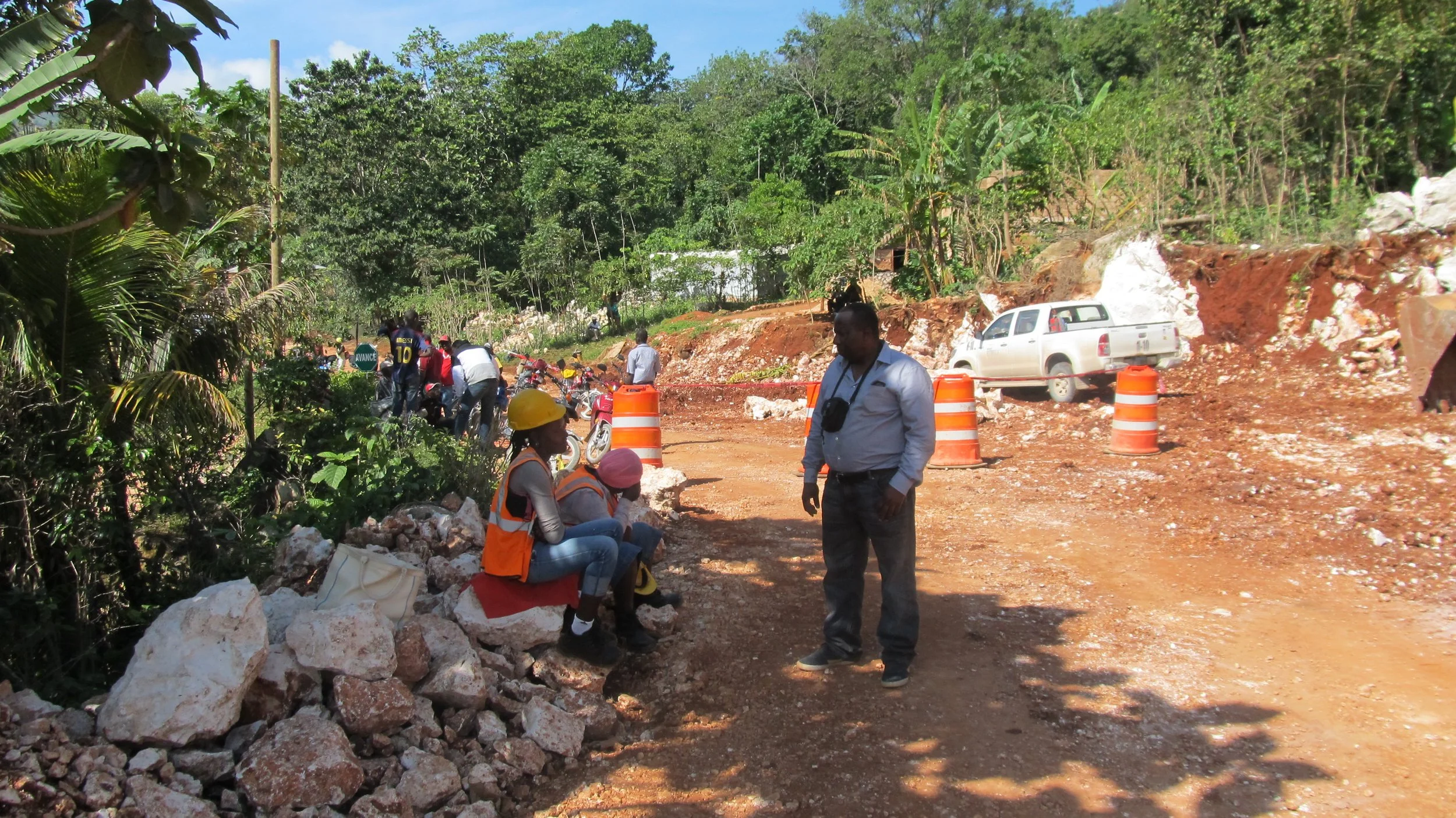 Prof. Ambrose Walemba and some people were working and sitting on a dirt road under orange traffic cones in a lush green area. Some are wearing safety vests and hard hats. A white pickup truck is parked in the background surrounded by trees. Haiti.