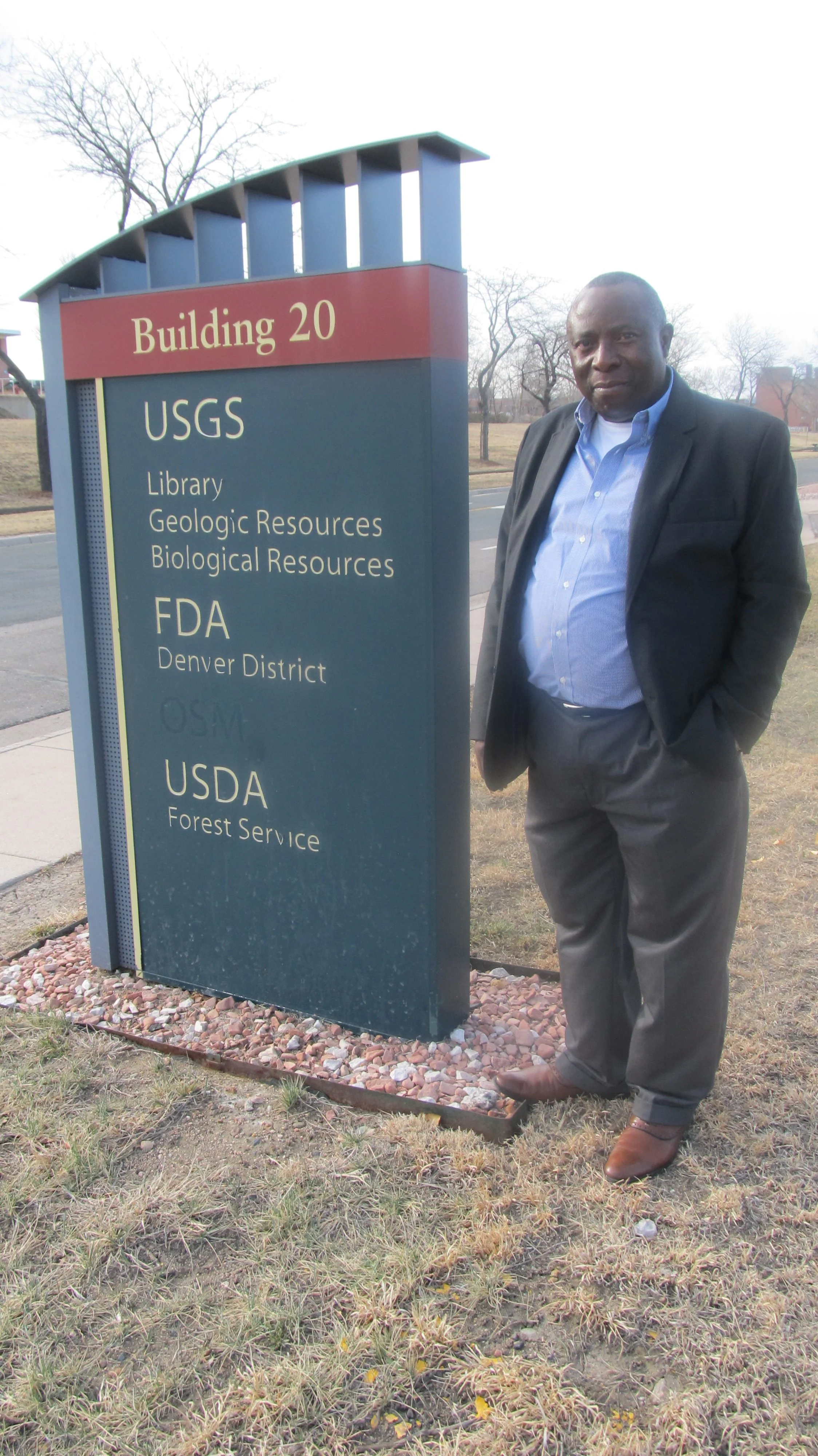 Prof. Ambrose Walemba in a black suit and blue shirt is standing next to a sign for Building 20 with USGS, FDA, and USDA listed on it outside with trees and a sidewalk in the background. Denver, Colorado, USA
