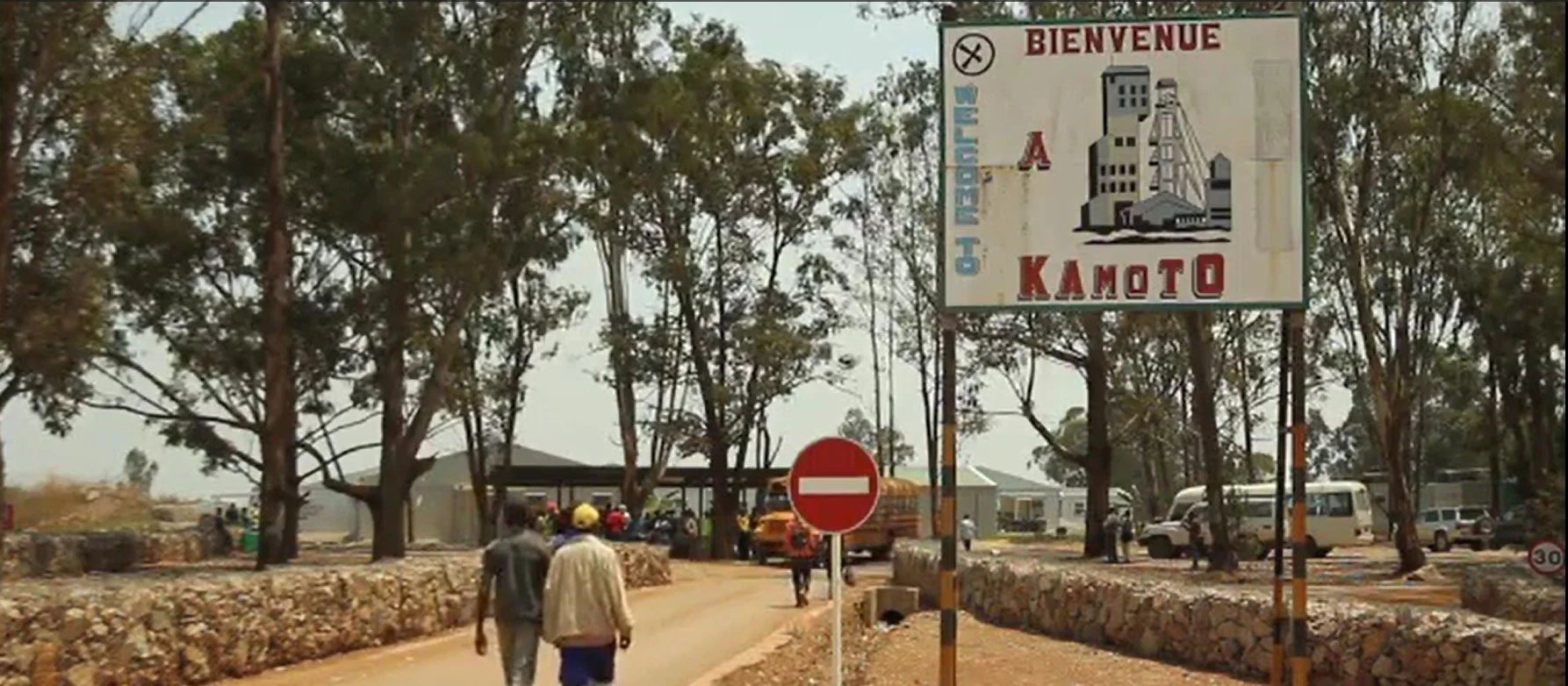 A roadside scene in Kamoto, with trees, a no entry traffic sign, and a large sign welcoming visitors to Kamoto, showing buildings and the words "BIENVENUE" and "KAMOTO." Several people are walking and vehicles are parked along the dirt road.