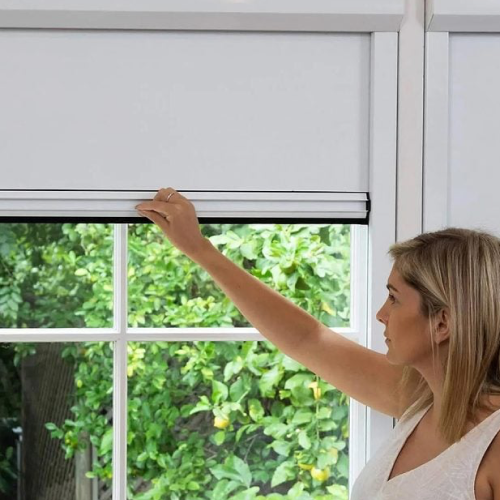 A woman with blonde hair has her arm up, closing or opening a white blackout shade with green bushes behind the window.