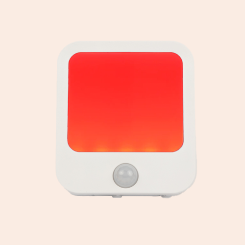 A white night light with rounded corners and a red light in its center. A motion-activated sensor is in the bottom center of the light. On a pink-ish background.
