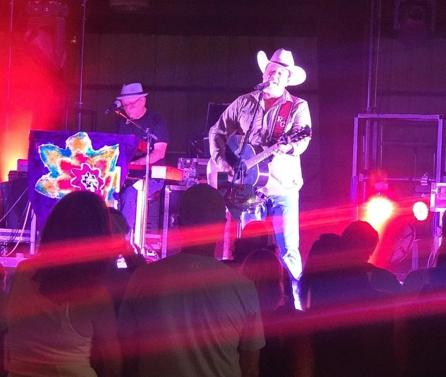 Musicians performing on stage with one playing guitar and wearing a cowboy hat, the other at keyboard with a colorful cloth or artwork in front, audience in foreground, colorful stage lighting.