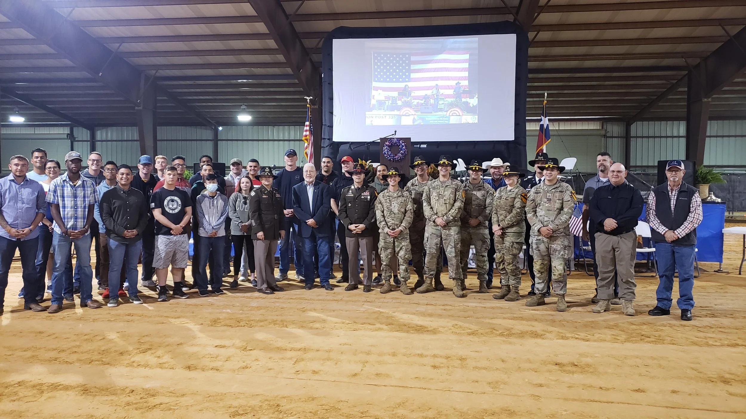A large group of people, including military personnel, civilians, and officials, standing together inside a covered pavilion with a large screen, American flags, and a wooden floor.