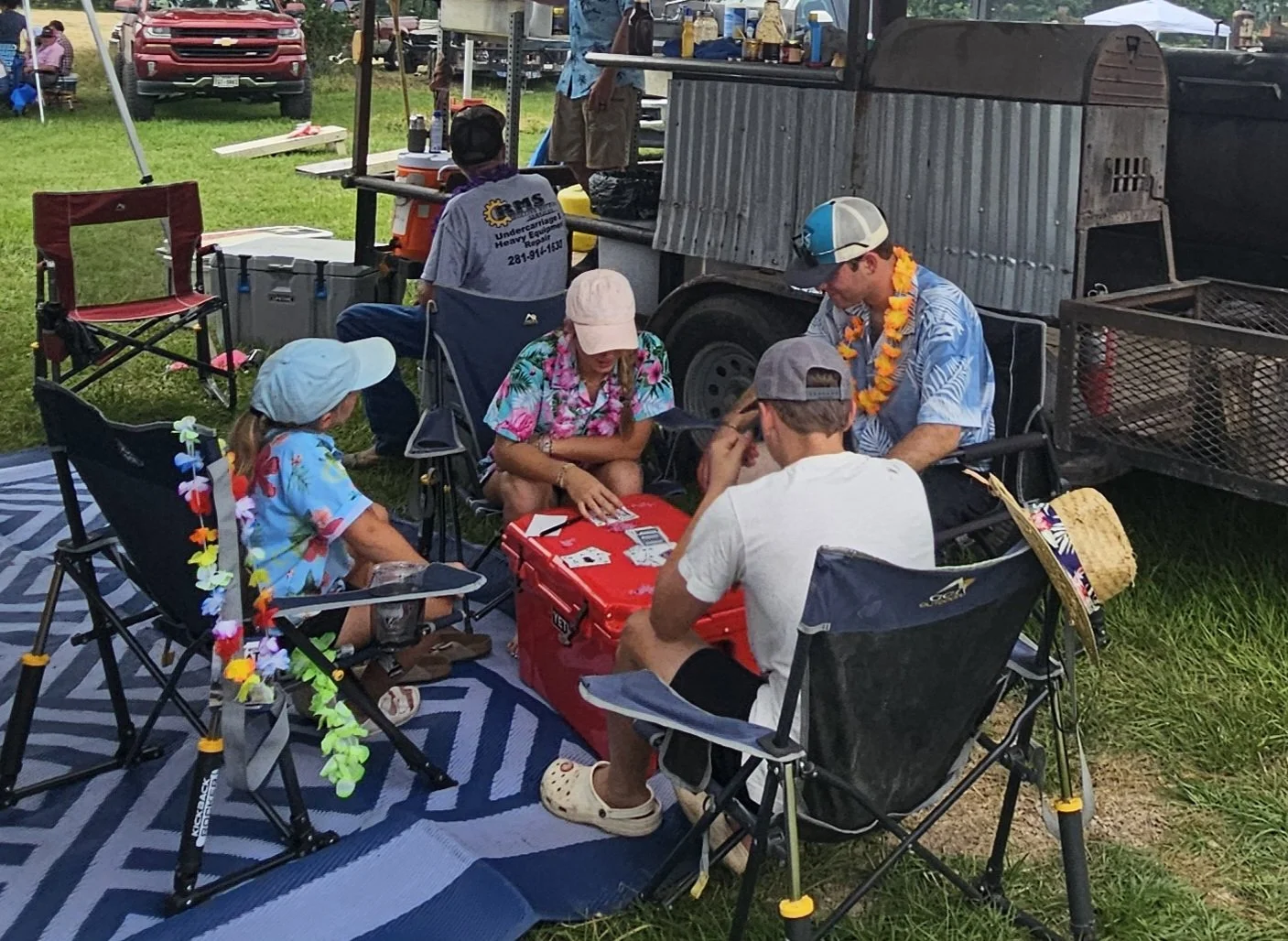 Group of people playing cards outdoors at a picnic or fair, sitting around a red cooler, with a food stand and grill in the background.